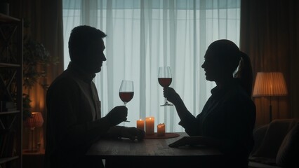 The couple spends the evening with red wine by candlelight, they say toasts, raising their glasses, talking. Dark silhouettes of a man and a woman in the living room at the table.
