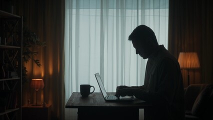 Side view of a dark silhouette of a man at a table in front of a laptop. A man is working on a...