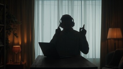 Dark silhouette of a man in wireless headphones at a table in front of a laptop. A man works on a...