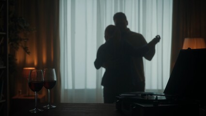 Vintage record player with a vinyl record and glasses with red wine on the table close up. A young couple is dancing a slow dance while enjoying a pleasant evening together. Silhouette. - obrazy, fototapety, plakaty