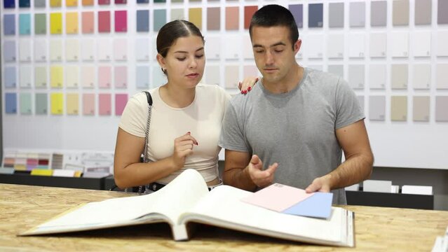 Positive young adult couple, woman and man, flipping through paint color catalog and discussing with interest future design of house under renovation in painting supply store