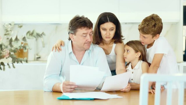 Upset father reads and looks through financial documents. Sad wife with children in the background. High quality 4k footage
