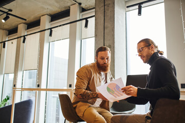 stylish entrepreneurs sitting near laptop in office with high tech interior and looking at paper with charts, businessman in eyeglasses and black turtleneck with bearded and tattooed colleague