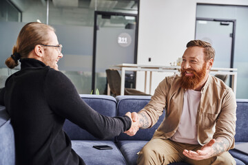 delighted, bearded and tattooed entrepreneur sitting on couch near smartphone and shaking hands...