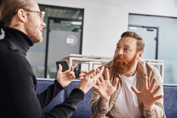 entrepreneurs gesturing and discussing startup project in modern office, bearded tattooed businessman with colleague in black turtleneck, business collaboration concept