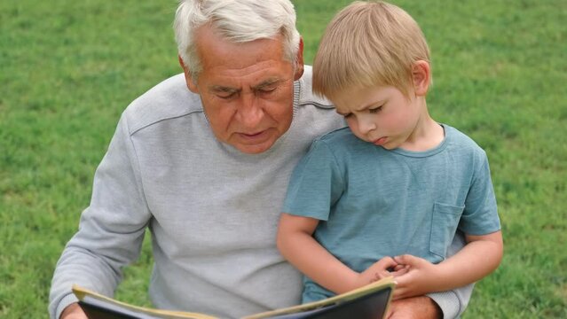 Happy grandfather reading book to curious grandson outdoors. Close up. Grandpa with grandchild spending time together. Family time comes in various forms. 4s year boy and senior man read story in park