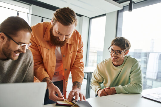 bearded and tattooed designer showing color samples to cheerful and stylish entrepreneurs in eyeglasses sitting near laptops in contemporary coworking environment, teamwork and business creativity