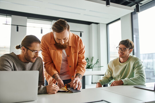 joyful businessman looking at bearded and tattooed designer showing color samples to colleague sitting near laptop at work desk in modern design studio, presentation of creative ideas