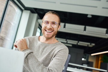 professional headshot of delighted businessman in stylish clothes and eyeglasses smiling at camera near laptop on blurred foreground in modern office, business productivity and success concept