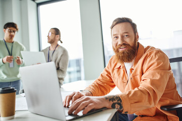 cheerful bearded and tattooed architect working on laptop near takeaway drink in paper cup and looking at camera while colleagues talking on blurred background in coworking design studio