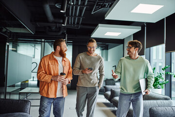 smiling entrepreneur in eyeglasses holding laptop and discussing startup with partners, walking with mobile phone and takeaway drink in lounge of modern office environment