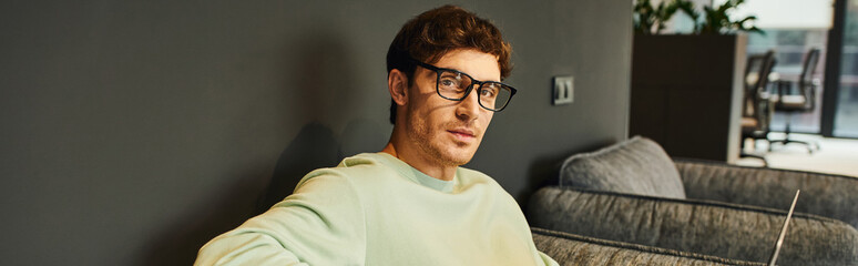 Obraz premium portrait of stylish businessman in casual clothes and eyeglasses looking at camera in contemporary office environment, professional headshot, confidence and success concept, banner