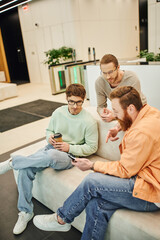 bearded businessmen pointing with finger and showing smartphone to colleagues in eyeglasses while sitting on couch in lounge of modern coworking office during coffee break