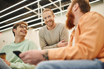 low angle view of bearded entrepreneur with mobile phone discussing possibility of business startup with cheerful colleagues in eyeglasses during coffee break in modern office on blurred foreground