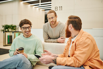 bearded man with mobile phone talking to smiling colleagues in eyeglasses while sitting on comfortable couch during coffee break in lounge of modern office, positive entrepreneurs discussing startup