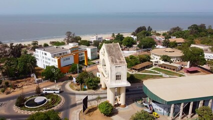 Aerial parallax shot of the Arch 22 memorial the triumphal arch of banjul city in the gambia with a view of the sea and a busy road with traffic at a roundabout and buildings on a beautiful beach