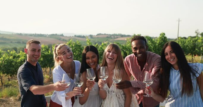 Happy adult friends cheering white wine while smiling on camera with vineyard in background - Multiracial people doing party at summer time in countryside resort