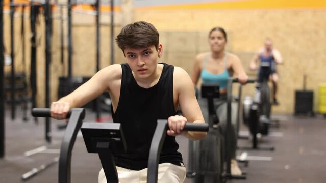 Dedicated active strong young man doing cardio workout on stationary bicycle as hard cross-training in gym indoors