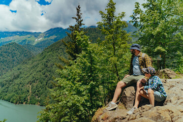 Hiking traveller caucasian bearded man with backpack sitting on rocks with son enjoying beautiful view of turquois blue Lake Ritsa in Abkhazia