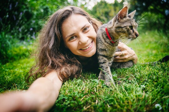 Happy young woman taking selfie with cute gray cat lying on green grass in summer garden