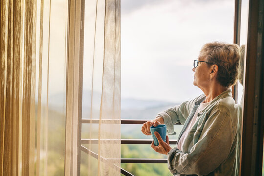 Beautiful senior elderly woman standing at the window overlooking the mountains and holding a mug of coffee in her hands - Powered by Adobe