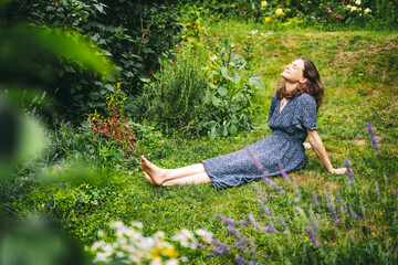 Happy young woman in dress relaxing and enjoying summer while sitting on green grass in her garden - 621853365