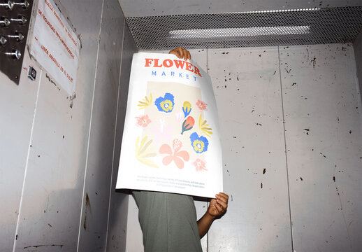 Mockup Of Woman In Elevator Holding Customizable A2 Poster In Front Of Face, Low Angle