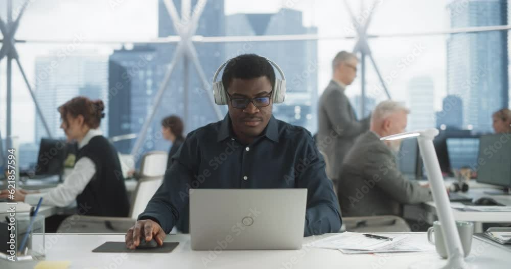 Poster Succesful Black Man Puts On Headphones And Starts Typing On Laptop Computer In Skyscraper Office In Downtown. Professional Male Software Developer Working On a New Project In Diverse Tech Startup.