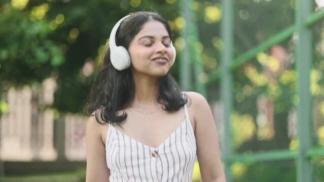 Portrait of pretty young indian woman with headphones listening to music and looking around outdoors Happy relaxed lady walking down and dancing the city street enjoying beautiful day alone