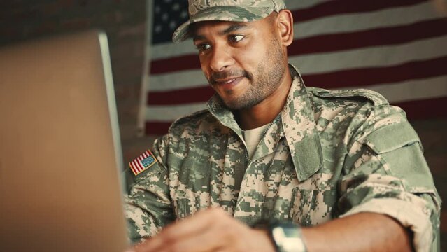 Soldier having a video call with his family from the military, he is using a laptop and sitting with an American flag behind him. Male US Marine catching up with his loved ones during his deployment.