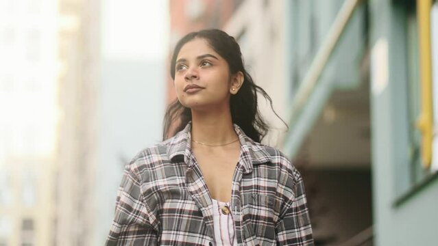 Charming young indian woman walking down the street with flying hair and looking around outdoors Happy relaxed lady walking on the city centre enjoying beautiful day	