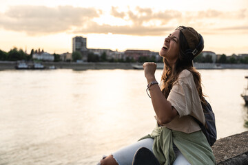 Happy teenage female tourist sitting on sea promenade of an old city in sunset. Young woman world traveler singing her favorite song on headset, making a break from walking through the city.