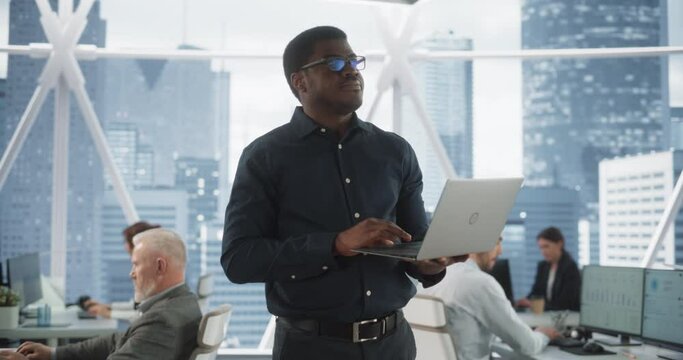 African American Financial Analyst Walking In Skyscraper Office With Diverse Employees In Downtown. Succeful Black Man Using Laptop Computer To Go Through Tasks And Business Mettings In Hedge Fund.