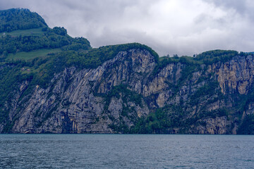 Obraz premium Scenic landscape with cliff and woodland at lakeshore of Lake Lucerne seen from passenger ship on Lake Lucerne on a cloudy spring day. Photo taken May 18t, 2023, Treib, Canton Uri, Switzerland.