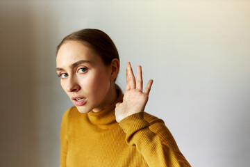 Portrait of concentrated anxious female eavesdropping holding head next to ear, listening carefully to strange sounds, feeling scared, standing against gray studio background with opened mouth