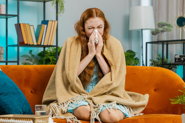 Young ill redhead woman suffering from cold or allergy sitting on sofa in living room at home. Sick girl wrapped in plaid blows sneezes wipes snot into paper napkin. Coronavirus quarantine, pandemic