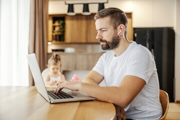 A focused remote worker is typing on a keyboard on a laptop while babysitting his daughter. - obrazy, fototapety, plakaty