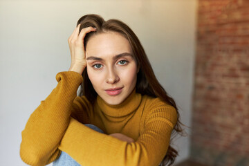 Tired female of 20s sitting against red brick wall with hands around knees, touching hair, feeling exhausted with lots of destroying thoughts in her head, looking at camera with smile and sad eyes