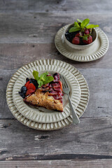 A piece of homemade summer galette with fruits, berries and mint in a decorative ceramic plate with a fork on a wooden table and a bowl with fresh strawberries and blueberries next to it. 