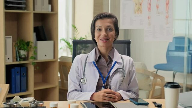 Portrait of smiling therapist in uniform sitting at desk at workplace and looking at camera