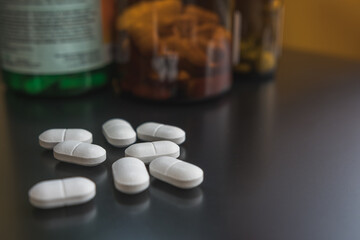 White pills on a black surface, selective focus, close up. Defocused medicine bottles in the background.
