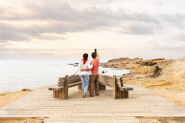 Couple on beach calling on phone taking selfie on wooden porch at sunset with sea in background