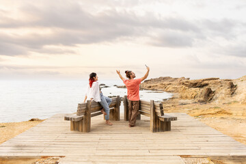Couple on beach calling on phone taking selfie on wooden porch at sunset with sea in background