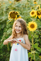 The cute little girl in a white dress with long beautiful hair laughing in a field of sunflowers and showing a heart. Portrait of a beautiful child enjoying nature at sunset. Little explorers.