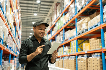 Warehouse worker wearing a hat and black shirt hands holding tablet check stock on tall shelves in...