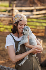 Portrait of young Caucasian female farmer in straw hat holding a little goat in her arms. Happy woman at the eco farm. Industrial production of goat milk dairy products. Cattle farming