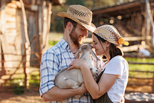 Portrait of young Caucasian happy farmer couple holding a little goat in their arms. Family business at ecological farm. Healthy food.Industrial production of goat milk dairy products. Cattle farming.