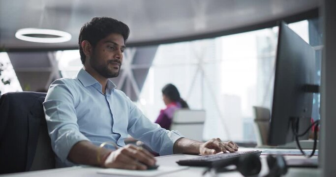 Portrait of a South Asian Financial Securities Trader Working on Desktop Computer in a Modern Office. Handsome Indian Accountant Bookkeeping Financial Affairs of a Business Venture