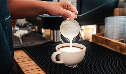 Hand of a barista pouring milk into coffee making cappuccino. Professional barista preparing coffee on the counter.