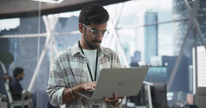 Indian Male Business Analyst Standing with Laptop Computer in a Modern Office. Successful Man Assessing Technological Risks And Developing Strategies For Company Growth and Expansion